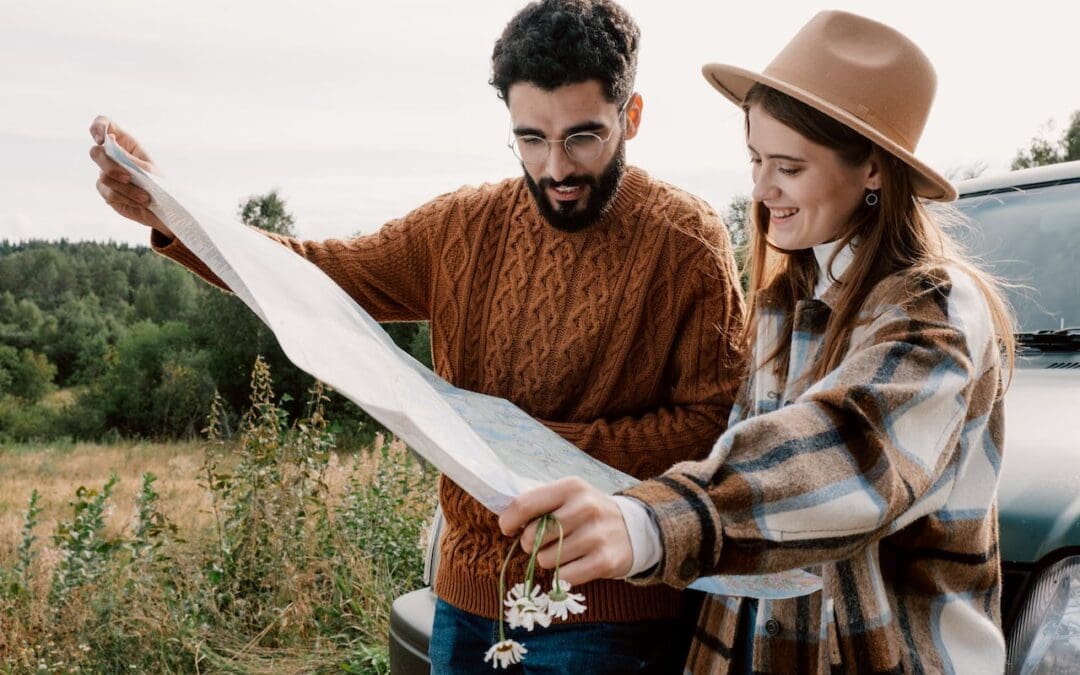 A young man with glasses and woman with a beige hat look at a map together as they stand outside of their car, parked next to a field.