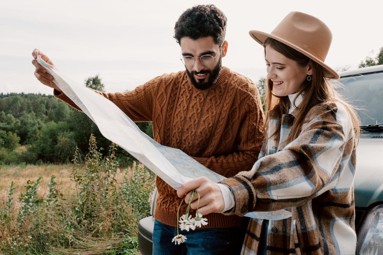 A young man with glasses and woman with a beige hat look at a map together as they stand outside of their car, parked next to a field.