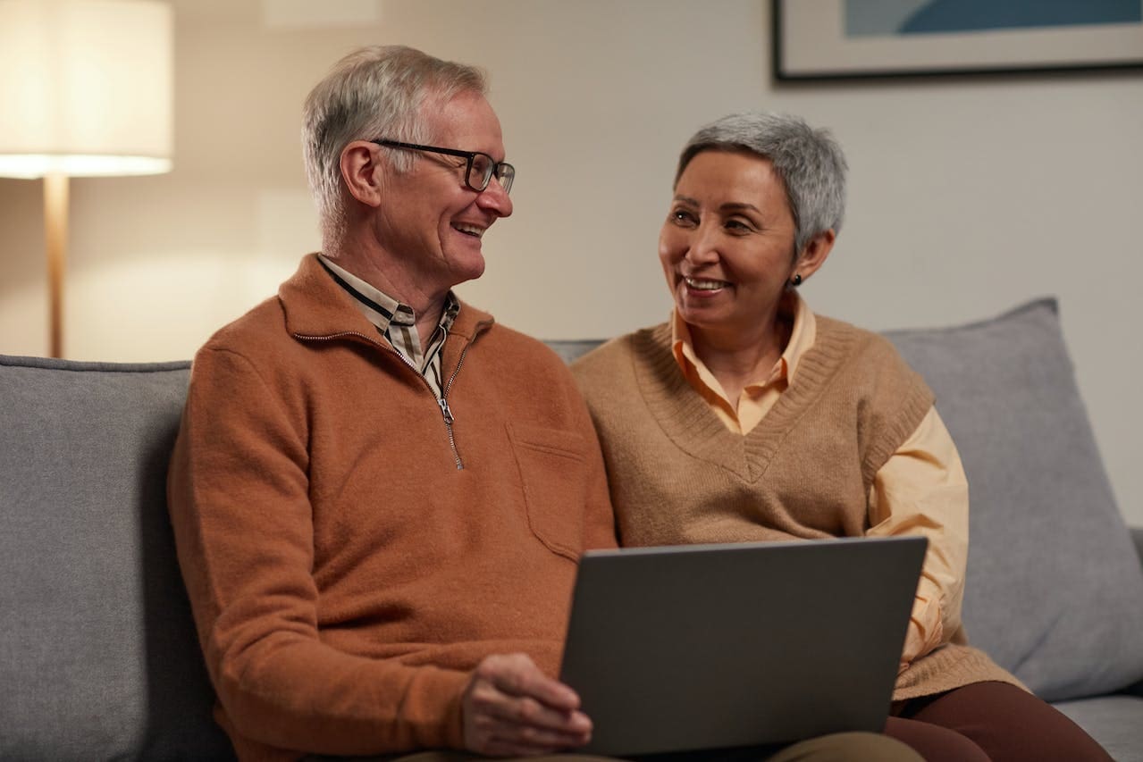 A couple in their 50s sit on a couch and smile at each other. The man, who wears glasses, holds a computer in his lap.