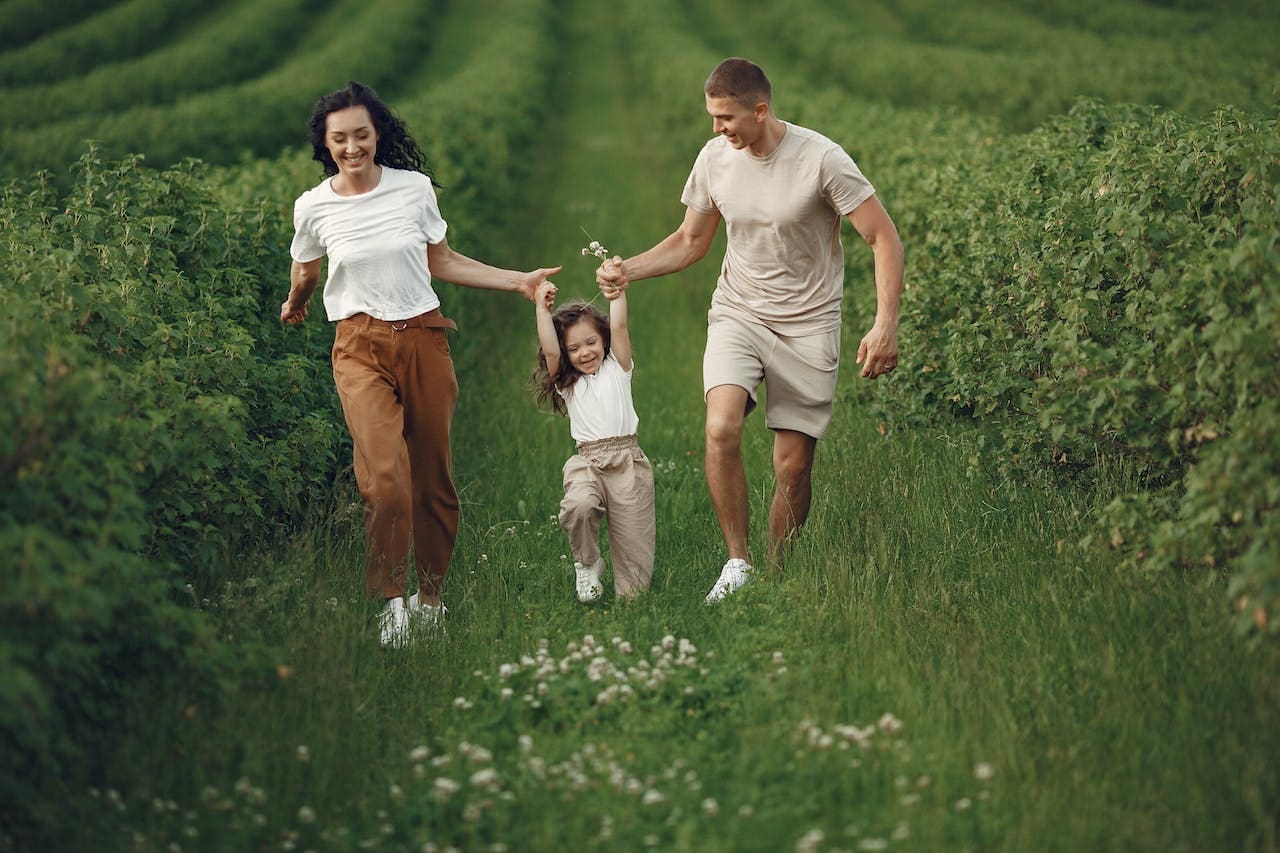 A smiling, young white couple run through rows of green plants, each holding the hand of a young girl as she swings between them and clutches white flowers.