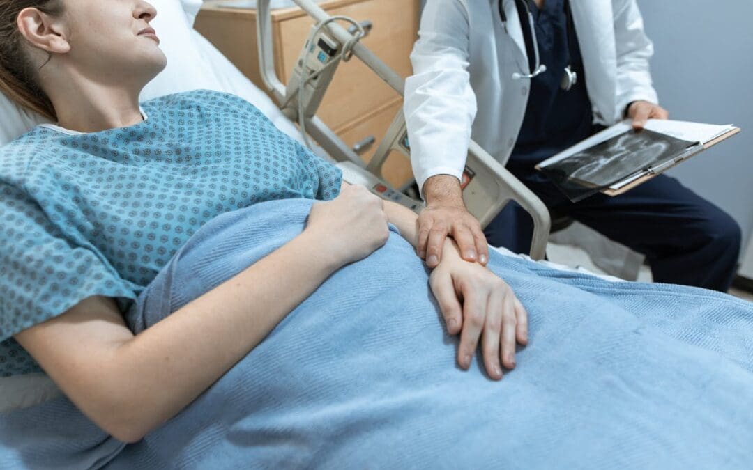 A doctor touches the arm of a female patient as she lays in a hospital bed.