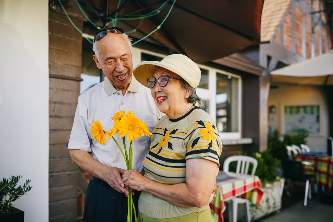 An older couple walks outside a cafe with a bouquet of bright yellow flowers.