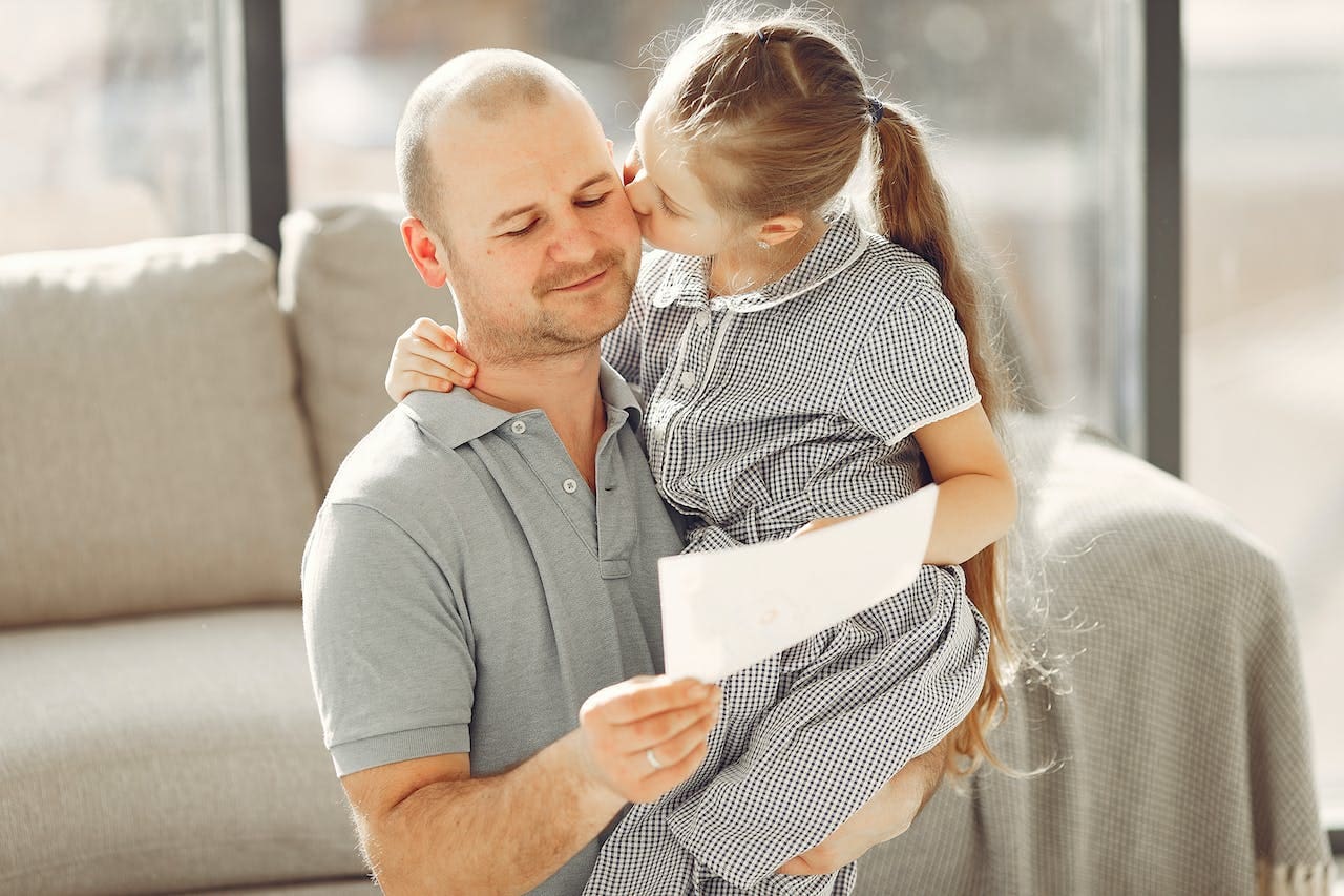 A dad with a low-cut beard holds his daughter, a girl with a long blonde ponytail, as she kisses his cheek. The man is absently reading a white paper while holding his daughter. They stand in front of a beige couch in a sunlit living room.