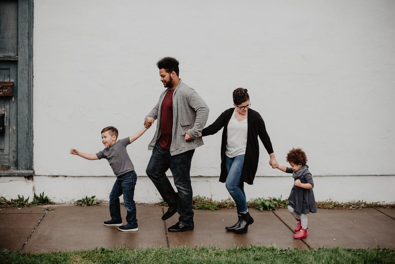 Photo by Emma Bauso via Pexels. A family of four walk down a sidewalk holding hands. A little boy leads the way, dragging his father, who’s wearing a gray sweater and a read shirt, forward. A smaller girl in a gray dress and red boots walks at the end of their line, holding her mother’s hand as she looks back to check on her.