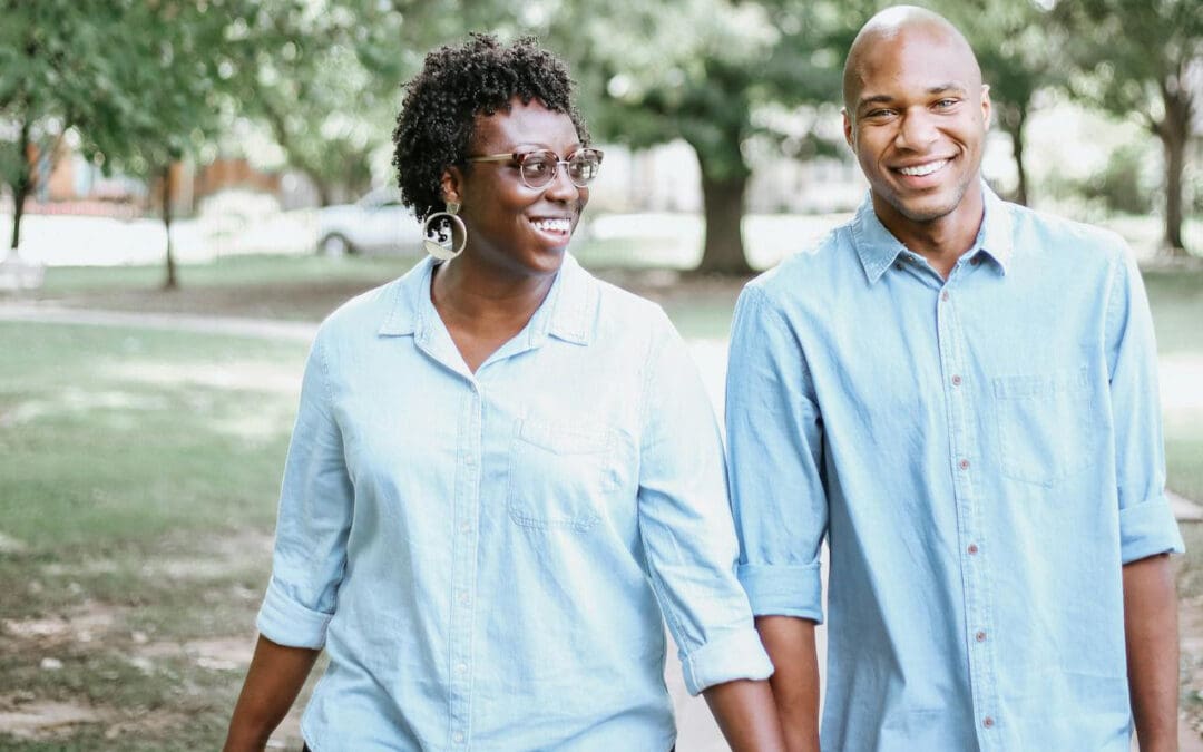 An older Black couple hold hands as they take a walk along a sidewalk under green trees. Both are wearing blue button down shirts. The woman, who’s wearing classes, looks at the man as they both smile.