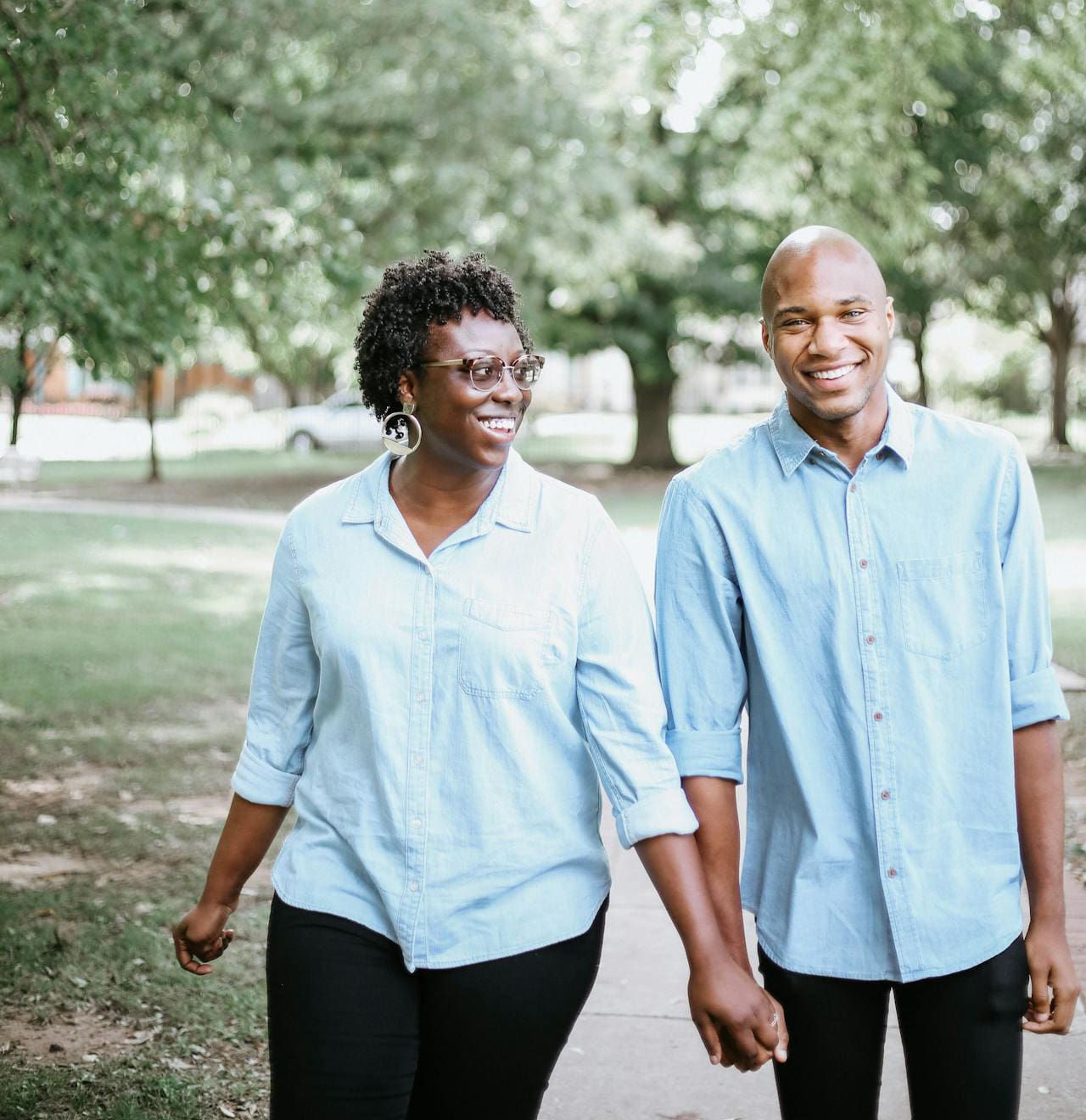 An older Black couple hold hands as they take a walk along a sidewalk under green trees. Both are wearing blue button down shirts. The woman, who’s wearing classes, looks at the man as they both smile.