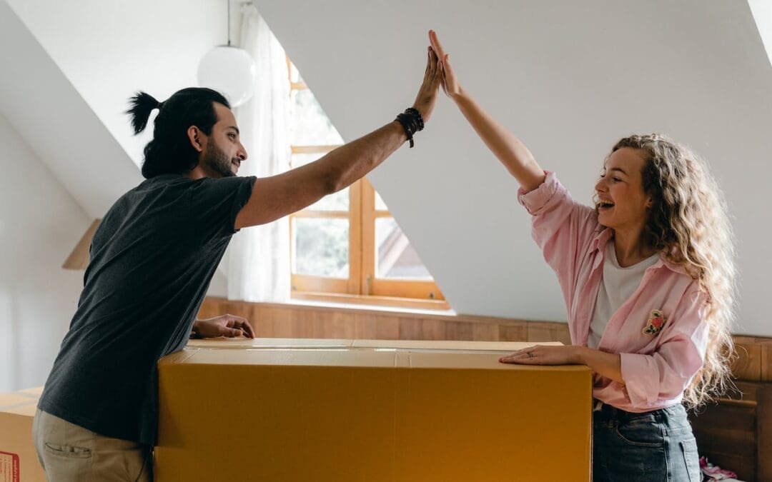 A young couple high fives over a large moving box. The white woman, on the right, is smiling and has long, curly light brown hair. She’s wearing a pink shirt and jeans. The white man, on the left, is wearing a black T-shirt and khaki pants. He has dark brown hair that is pulled back in a short ponytail and a beard.