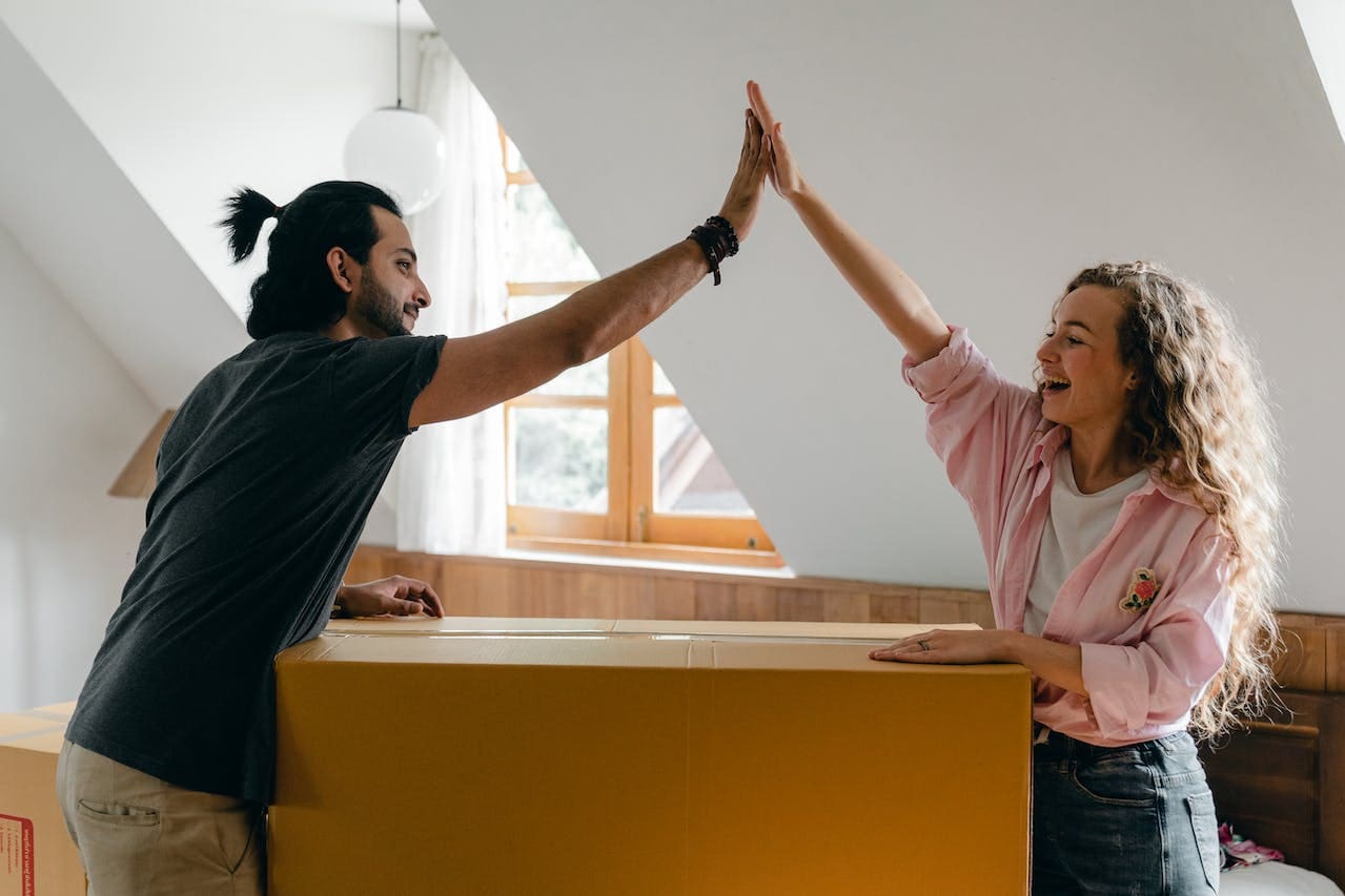 A young couple high fives over a large moving box. The white woman, on the right, is smiling and has long, curly light brown hair. She’s wearing a pink shirt and jeans. The white man, on the left, is wearing a black T-shirt and khaki pants. He has dark brown hair that is pulled back in a short ponytail and a beard.