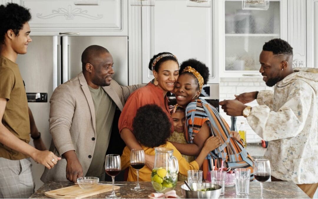 A large Black family goes in for a group hug in a modern-looking kitchen. Two younger girls are squished in between a mother and an older adult sister, who are both wearing headbands. Two young men on each side of the group are taller than the dad, who’s wearing a suit jacket. On the counter in front of them is a pitch of lemons and limes in water and an assortment of half-filled wine glasses.