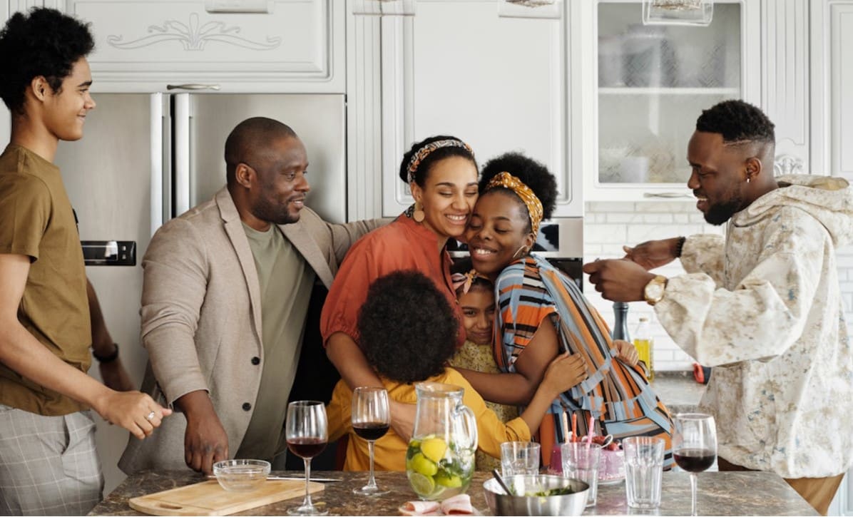 A large Black family goes in for a group hug in a modern-looking kitchen. Two younger girls are squished in between a mother and an older adult sister, who are both wearing headbands. Two young men on each side of the group are taller than the dad, who’s wearing a suit jacket. On the counter in front of them is a pitch of lemons and limes in water and an assortment of half-filled wine glasses.