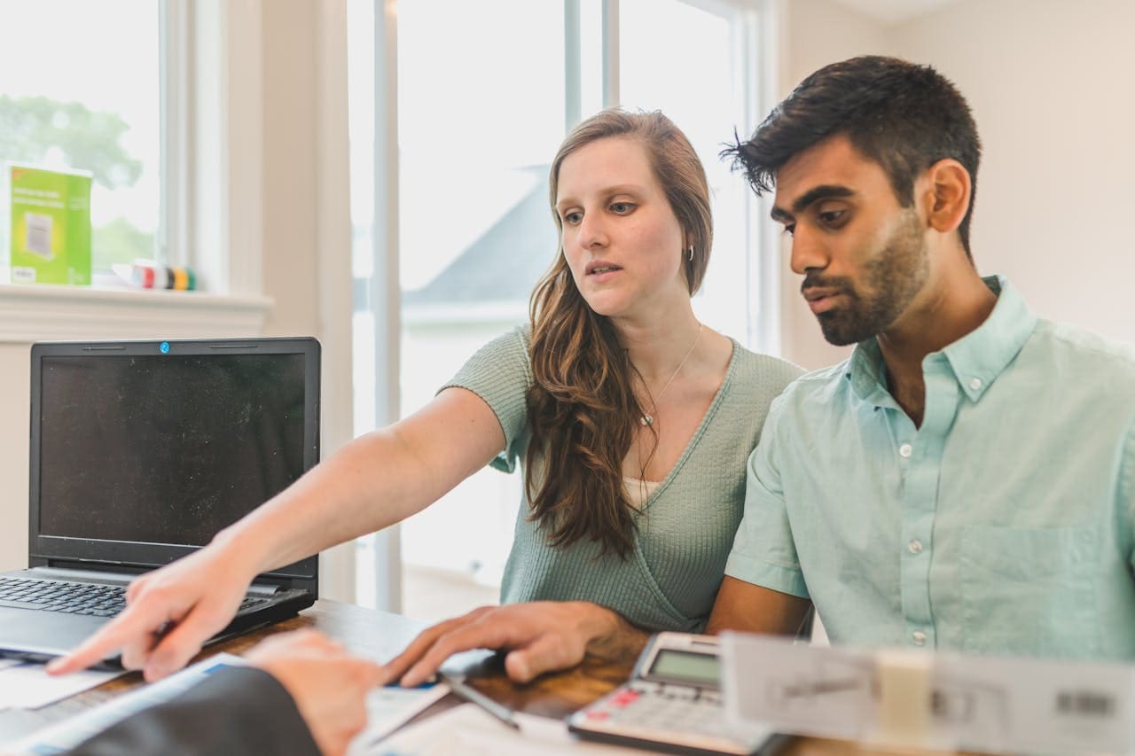 A white woman with long, brown hair points to a document in front of a man in a suit, who isn’t pictured. She’s seated by a dark-skinned man with a close-cut beard at a desk. They’re both wearing light green and look focused.