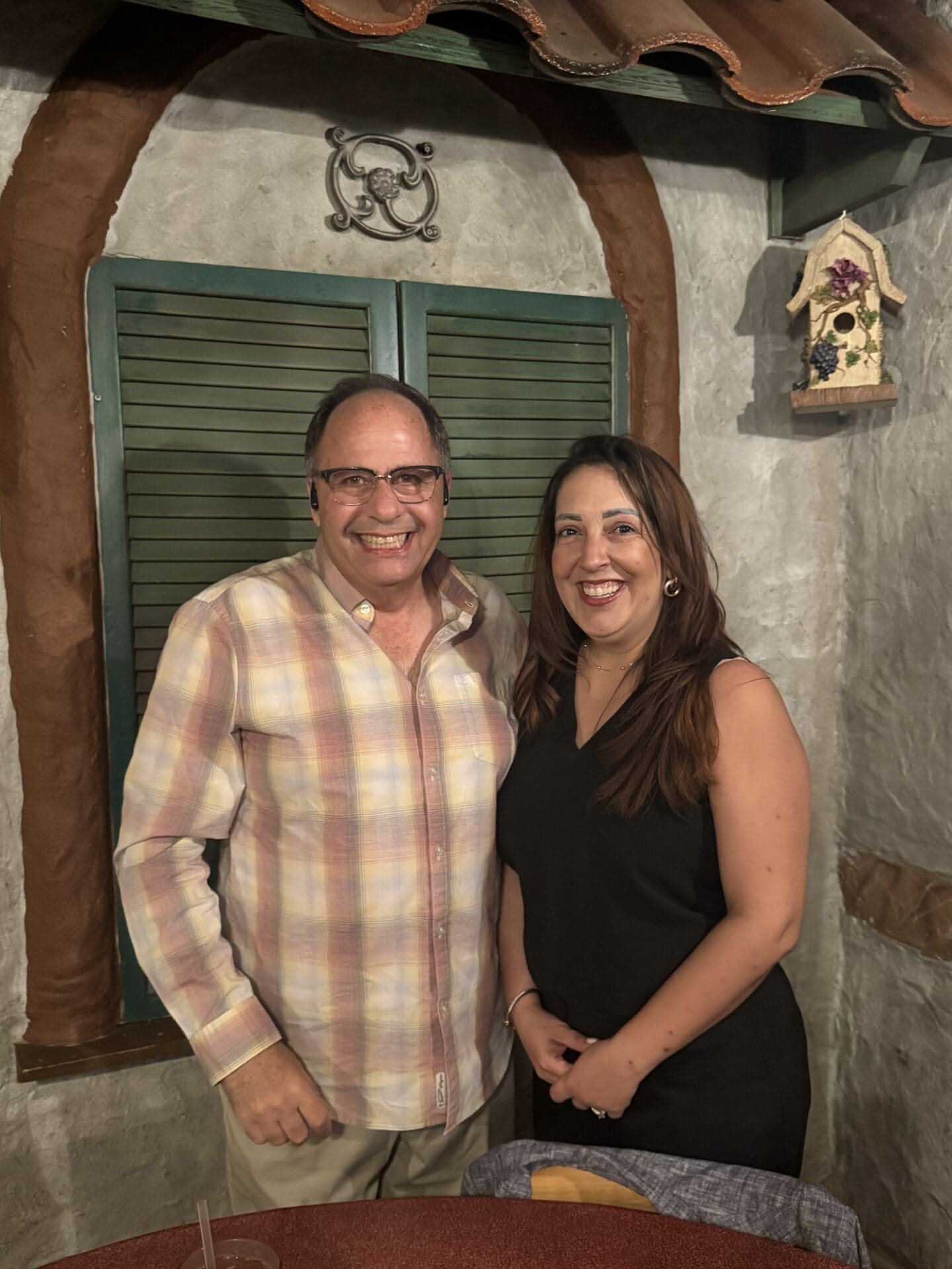A white man with glasses and a plaid shirt poses next to a Hispanic woman with dark hair and a gray suit. The wall behind them has a patch of exposed brick and a closed, green shutter.