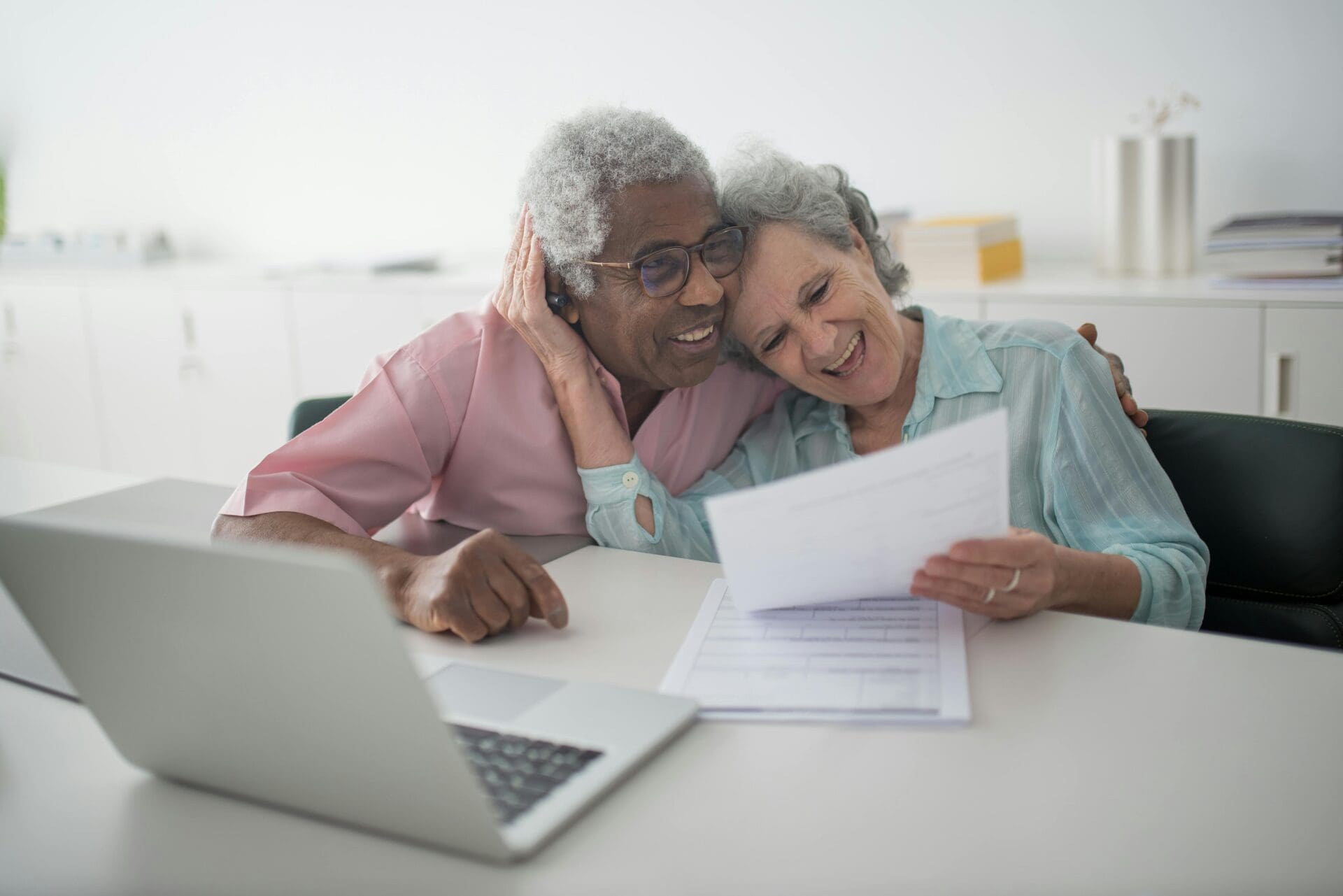 An older couple sitting down at a kitchen table, hugging and smiling looking over papers and a computer.
